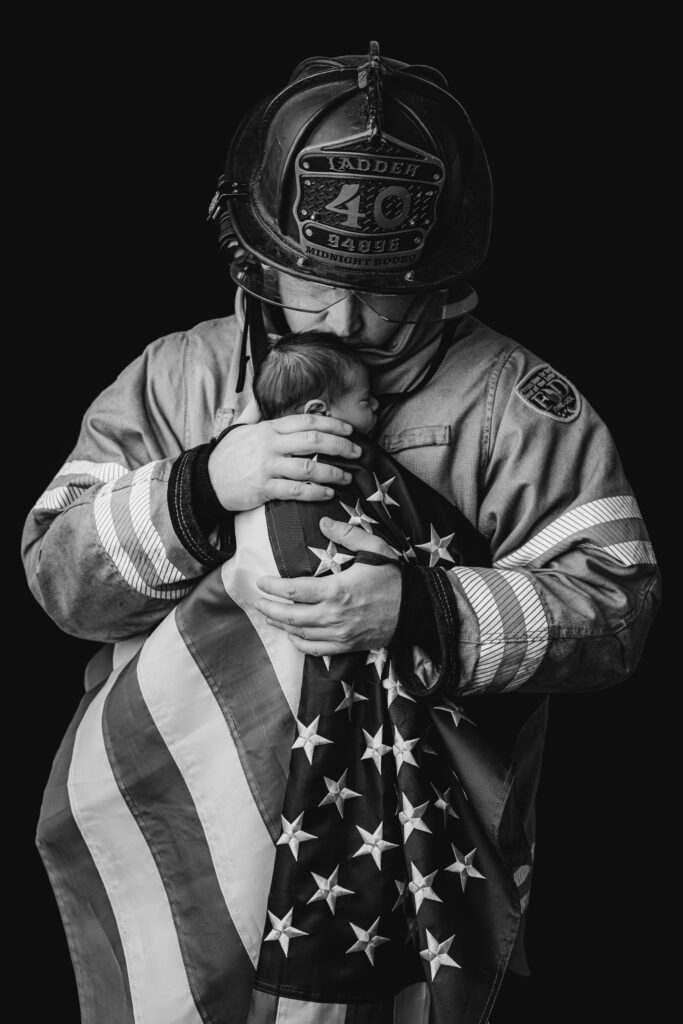 Black and white portrait of a Charlotte firefighter father in full turnout gear holding his newborn son wrapped in an American flag at NicSo Studio in Waxhaw, NC