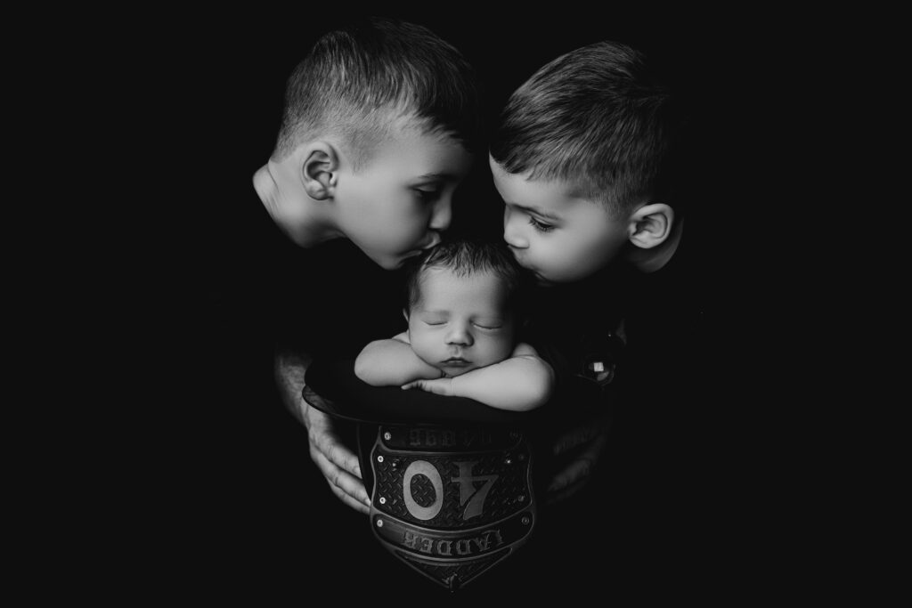 Black and white image of two big brothers kissing their newborn baby brother nestled inside a firefighter helmet during a Charlotte newborn session at NicSo Studio