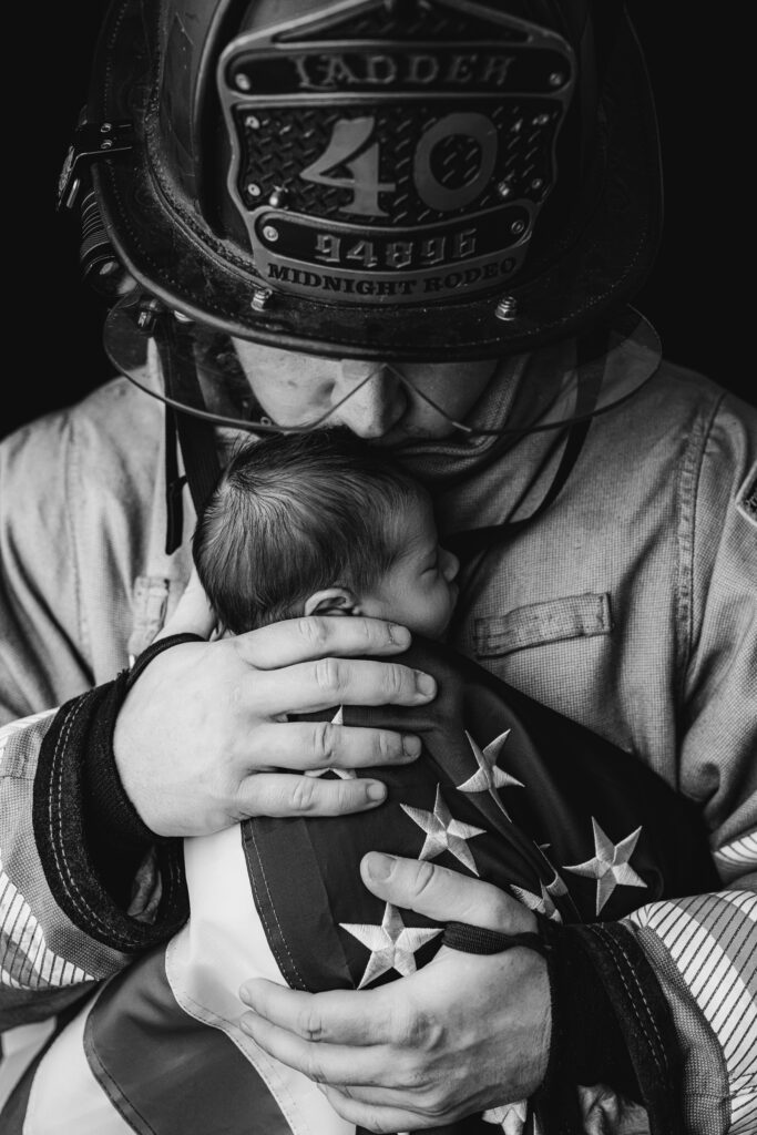 Close up black and white image of a firefighter father kissing his newborn baby's head while holding him wrapped in an American flag during a Charlotte newborn session