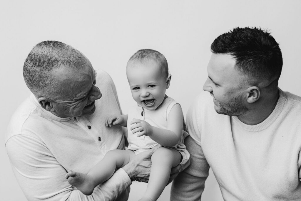 Black and white portrait of grandfather and father laughing with baby boy during extended family cake smash session
