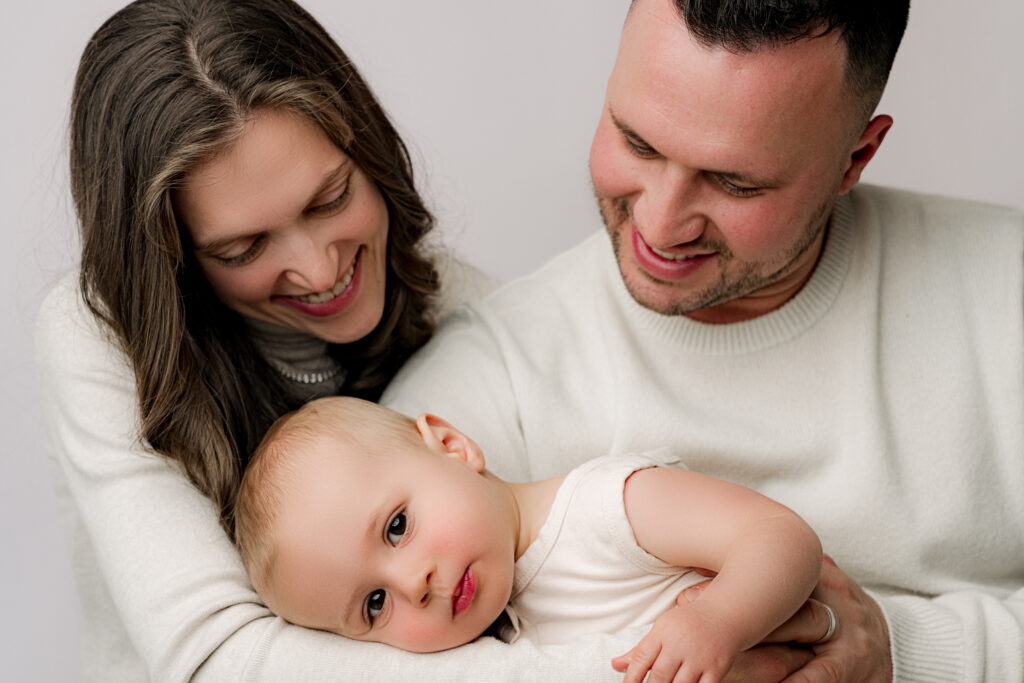 Mom and dad smiling down at baby boy laying in their arms during one year family portrait session