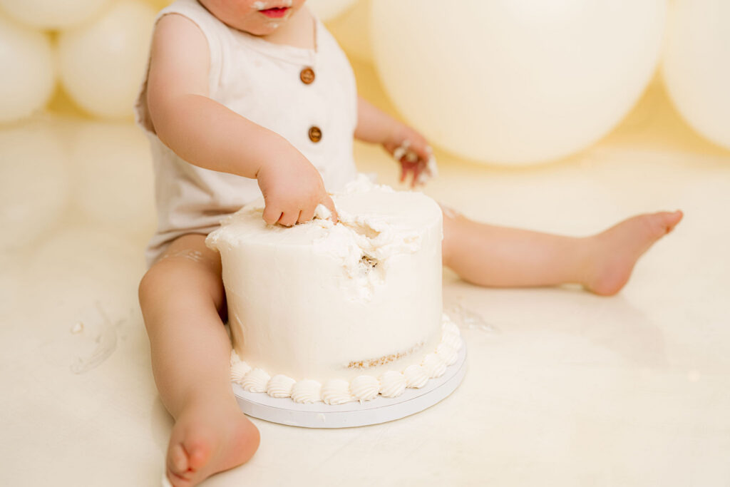 Baby boy reaching into smash cake during first birthday session at NicSo Studio near Charlotte NC with cream balloon backdrop