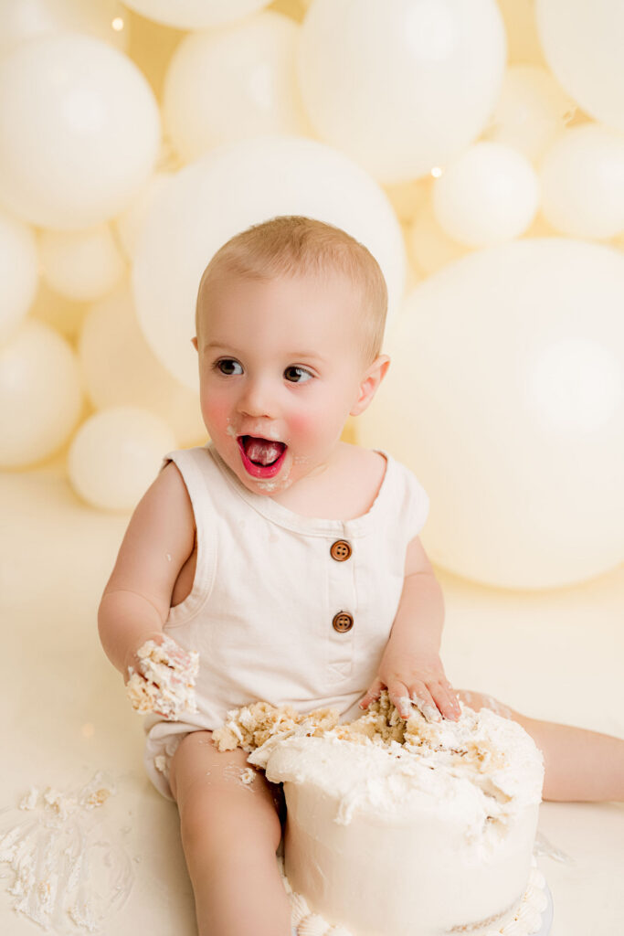 One year old baby boy standing at smash cake with cream balloon backdrop at NicSo Studio near Charlotte NC