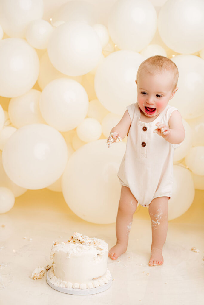 Baby boy standing smiling big looking down towards cake at first birthday smash cake in front of cream balloon wall