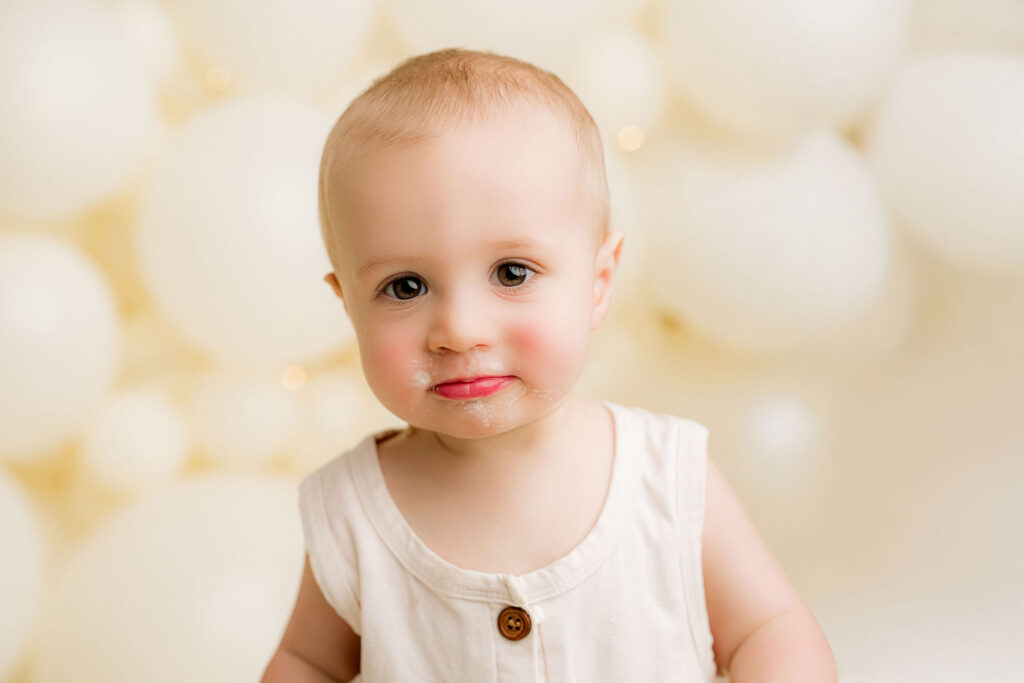 One year old baby boy with frosting on his lip smiling at camera during cake smash session at NicSo Studio in Waxhaw NC with cream balloon backdrop